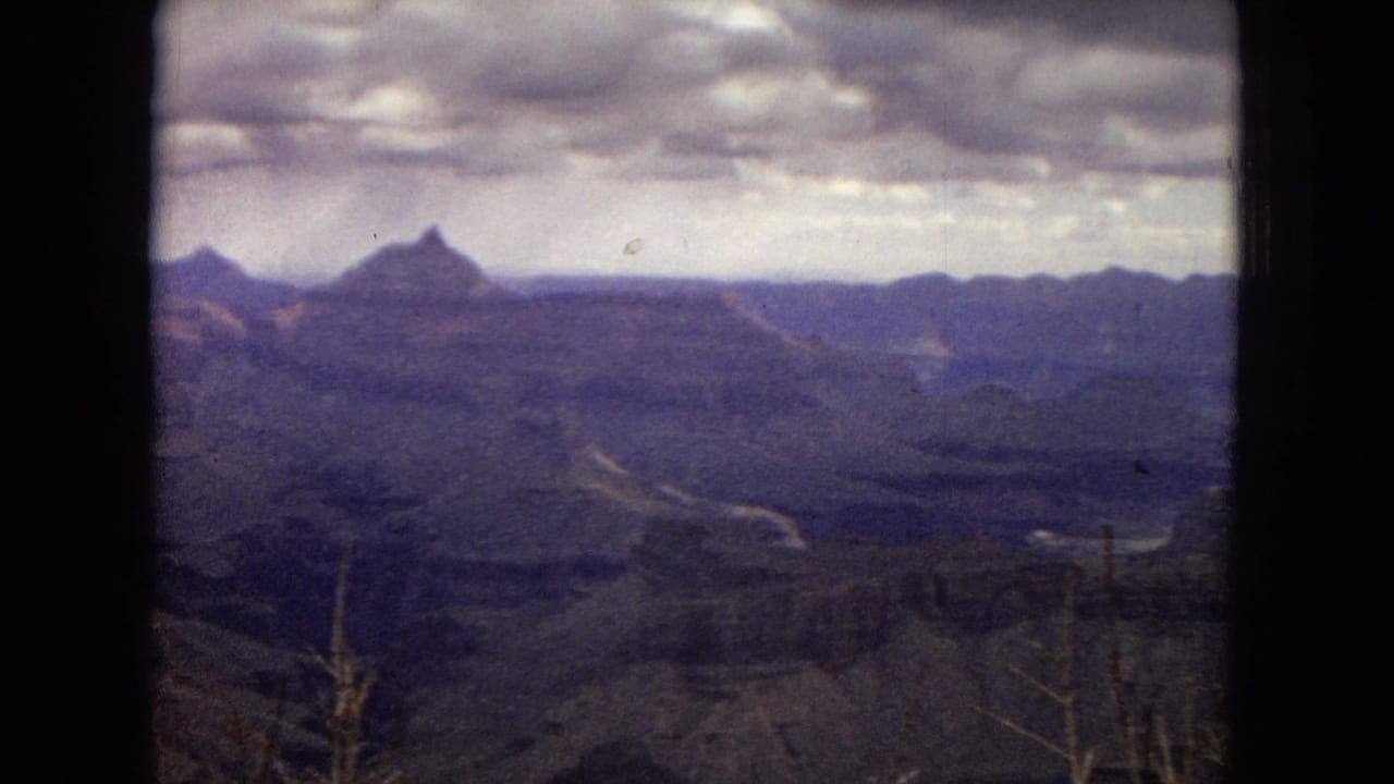 1967 Arizona 8mm frame showing a broad Grand Canyon view beneath a heavy overcast sky, with layered ridges fading toward the horizon and sparse shrubs in the foreground.