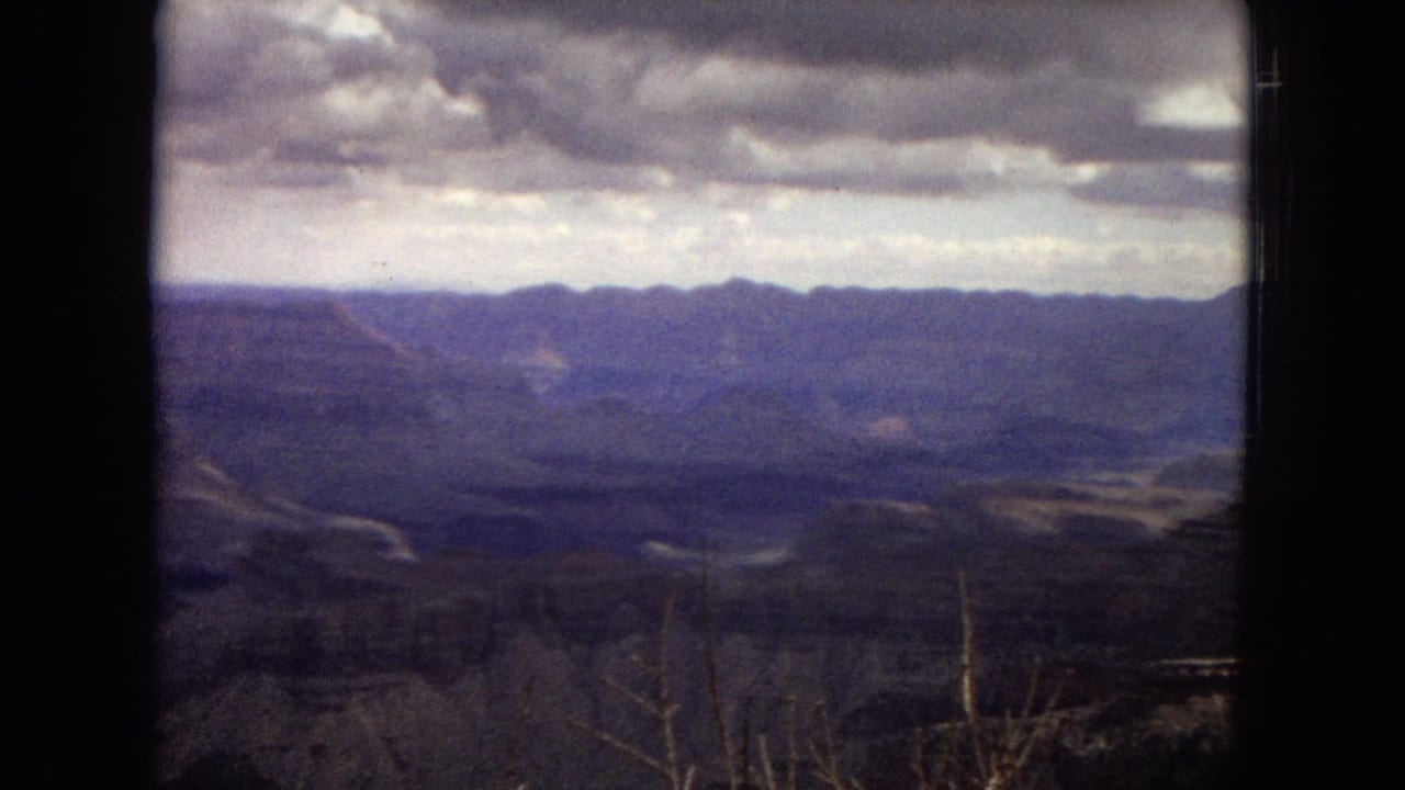1967 Arizona 8mm frame looking across the Grand Canyon toward a distant ridgeline under broken storm clouds, framed by the dark edge of the film scan.