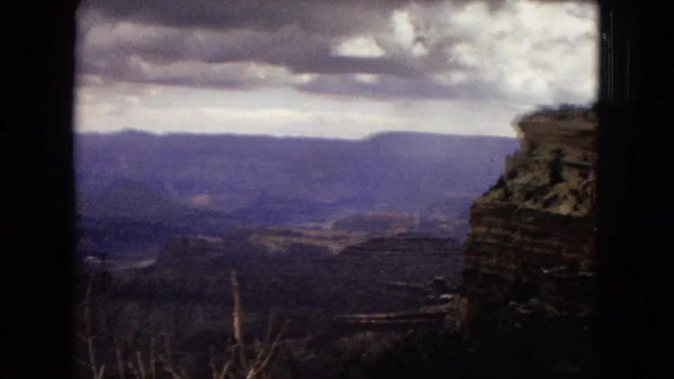 1967 Arizona 8mm frame from a canyon overlook, with a rocky ledge at the right edge, layered canyon walls below, and low clouds across the sky.