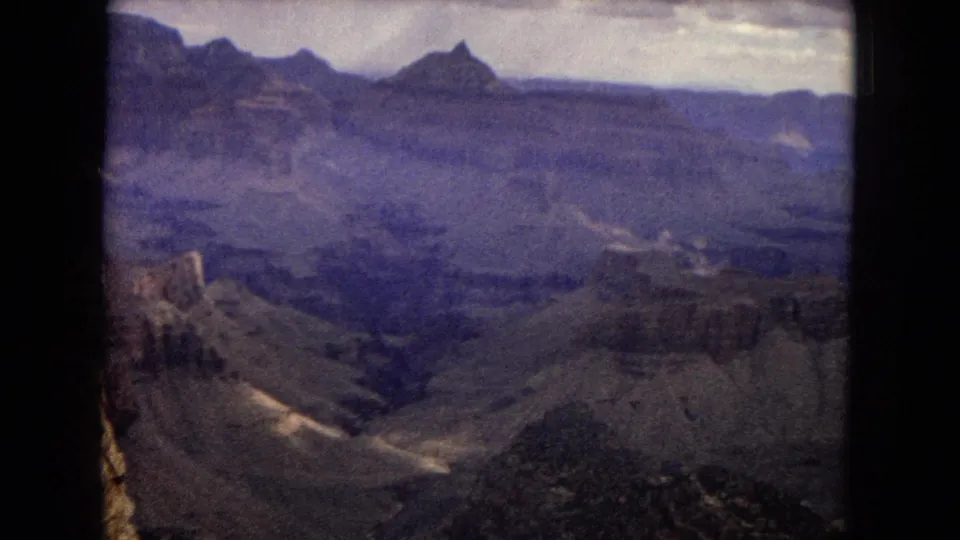 1967 Arizona 8mm frame showing a deeper Grand Canyon ravine with layered rock formations, distant pointed peaks, muted color, and a clouded sky.