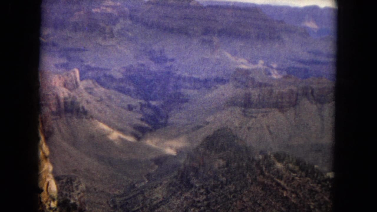 1967 8mm home-movie view of the Grand Canyon in Arizona, showing layered canyon walls, a wooded ridge, sparse brush, and dark film-frame edges.