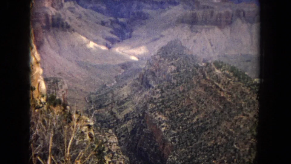 1967 Arizona Grand Canyon view with broad rock formations, forested slopes, pale snow patches, and a deep canyon basin below.