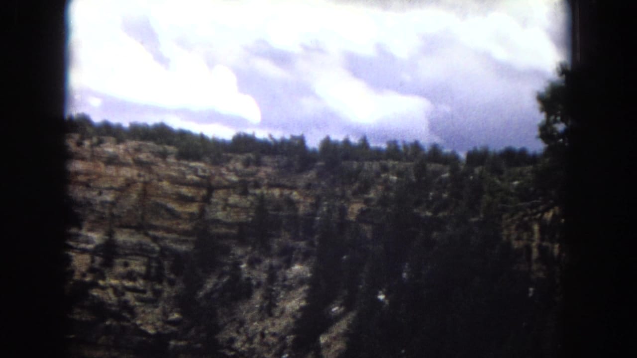 1967 Arizona 8mm frame with a darker view of layered canyon walls, ridge-top trees, and bright clouds above the overlook.