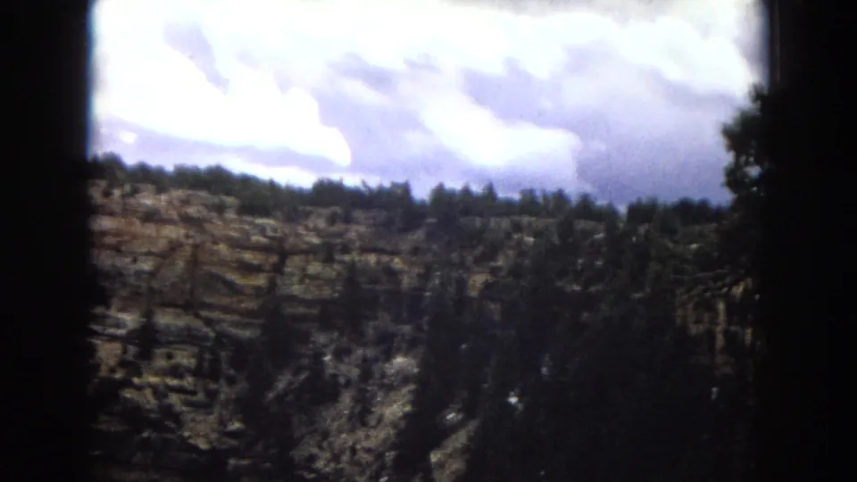 1967 Arizona 8mm frame with a darker view of layered canyon walls, ridge-top trees, and bright clouds above the overlook.