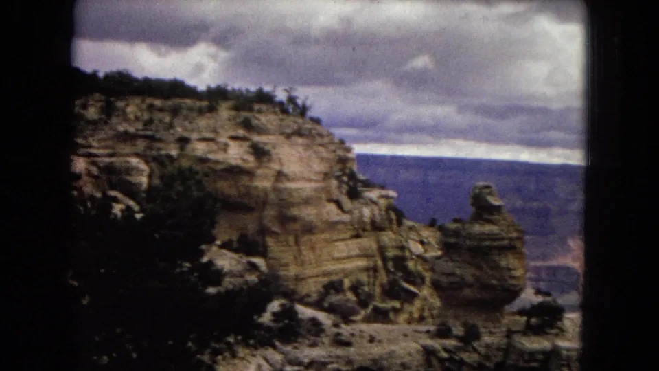 1967 Arizona home-movie view of a rock spire and canyon ledge with distant blue-toned canyon walls beneath a heavy cloud ceiling.