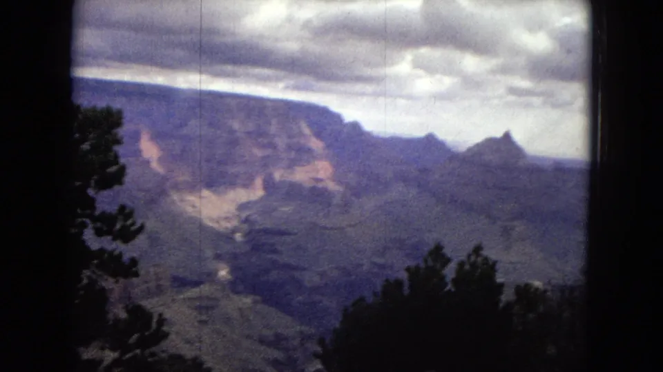1967 8mm view across the Grand Canyon, with foreground trees, broad shadowed canyon floor, layered cliffs, and heavy clouds.
