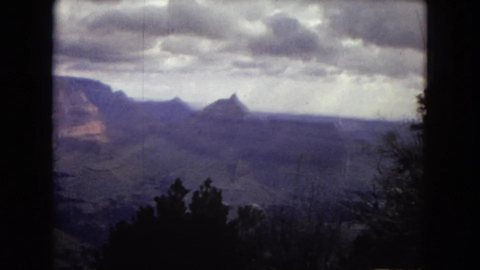 1967 Arizona home-movie frame of distant Grand Canyon formations, with silhouetted trees in the foreground and a cloudy sky above the canyon rim.