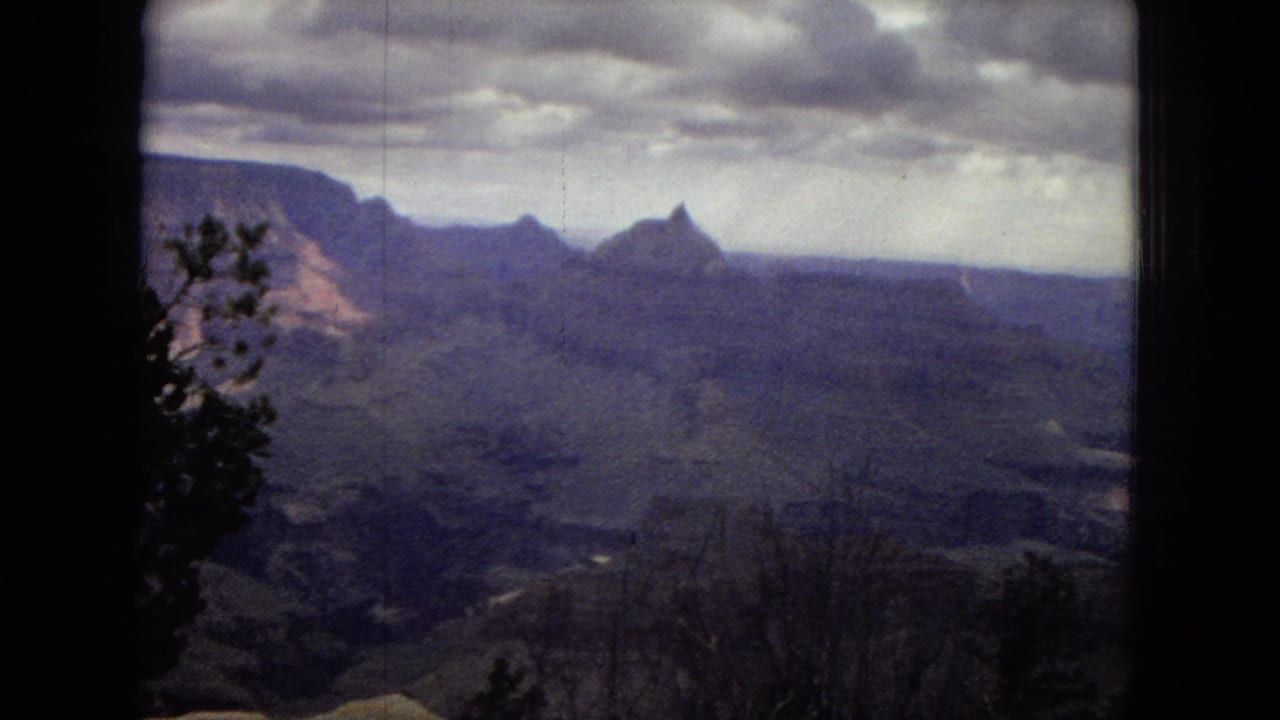 1967 8mm view of the Grand Canyon with layered canyon formations, distant ridges, heavy clouds, and silhouetted foreground trees.
