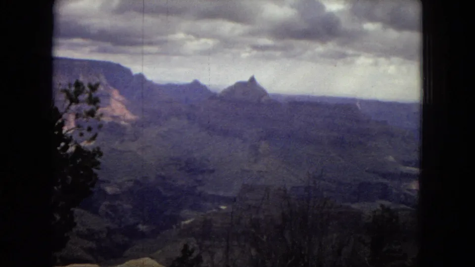 1967 8mm view of the Grand Canyon with layered canyon formations, distant ridges, heavy clouds, and silhouetted foreground trees.