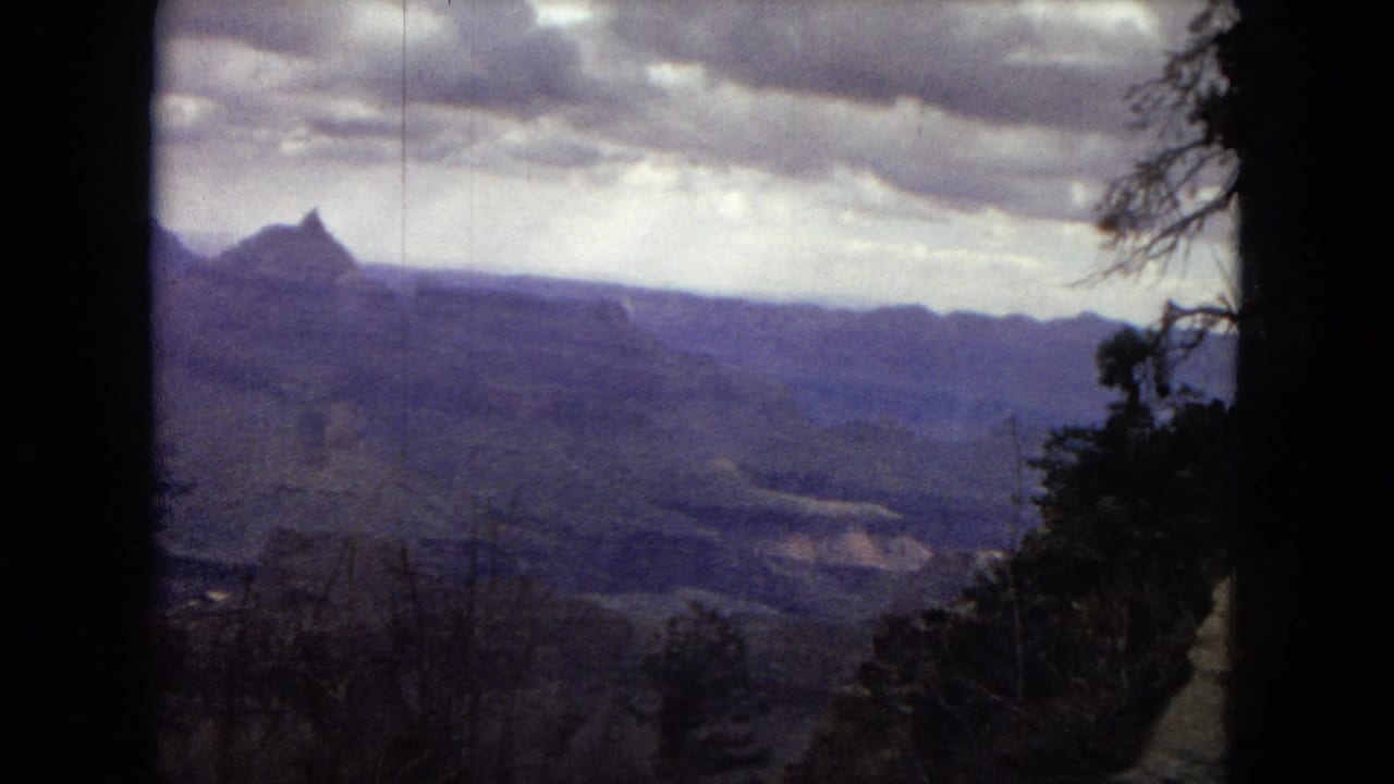 1967 8mm frame showing a broad Grand Canyon overlook, distant pointed ridge, layered valleys, cloudy sky, and dark foreground tree silhouettes.