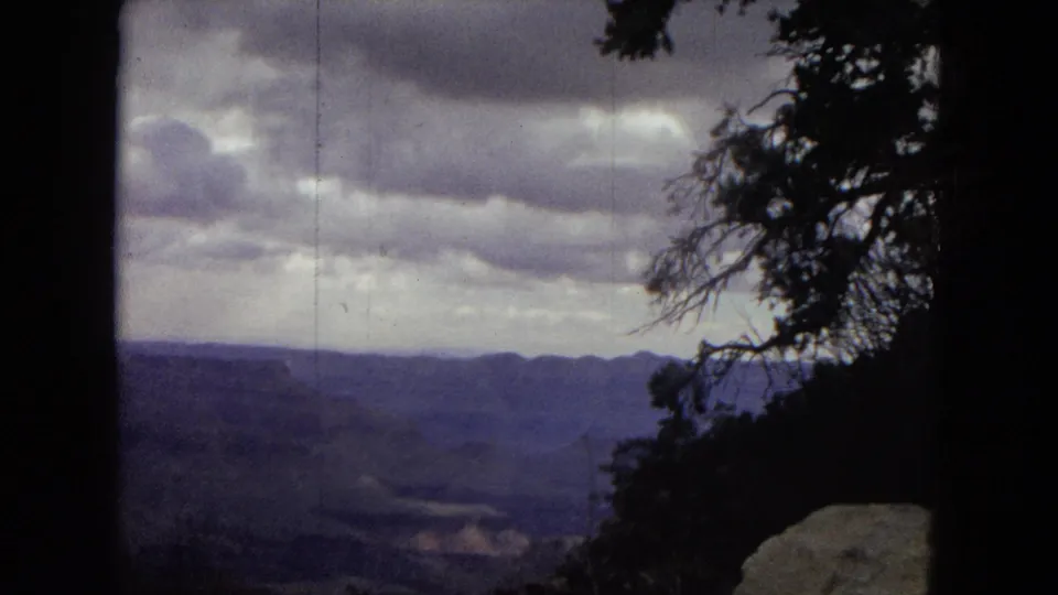 1967 8mm home-movie view across the Grand Canyon horizon, with heavy clouds, shadowed canyon layers, and a dark tree silhouette along the right side.