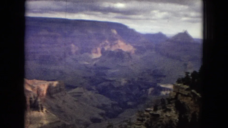 1967 8mm frame of the Grand Canyon showing layered cliffs, a rocky overlook edge, distant ridges, dark foreground vegetation, and a cloud-filled sky.