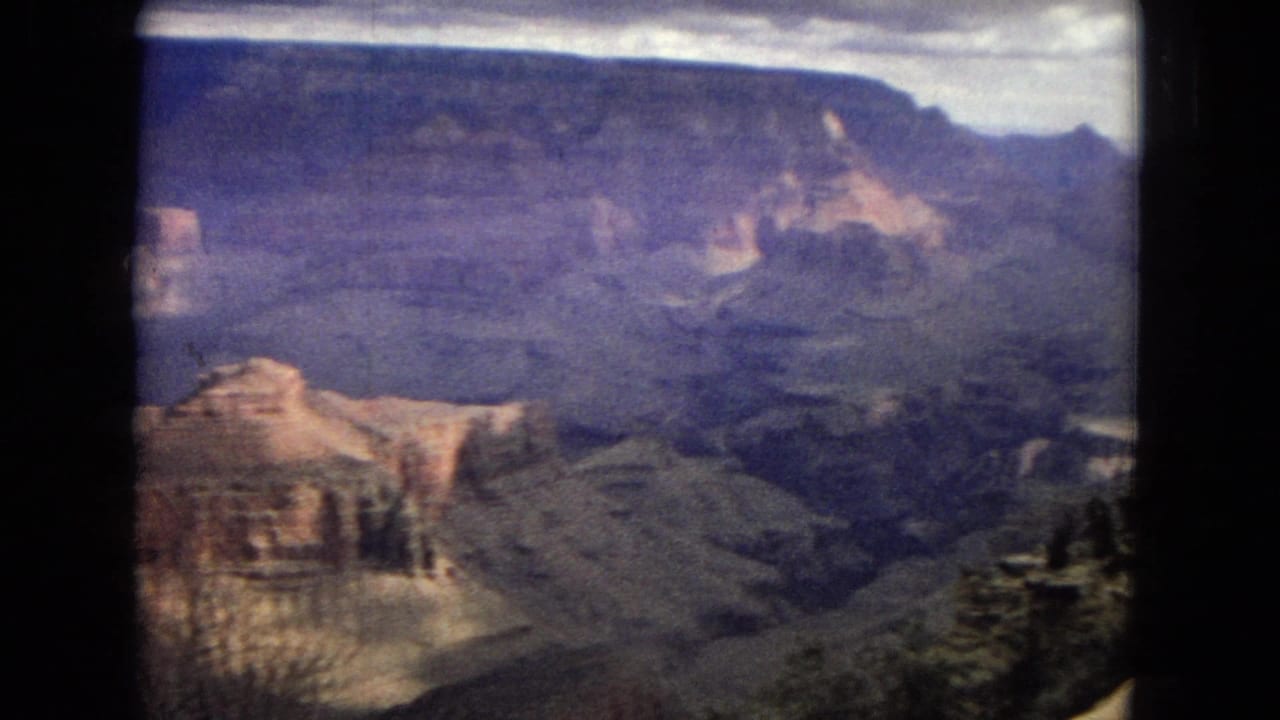 1967 8mm home-movie frame showing a wide Grand Canyon panorama with layered rock walls, deep ravines, and soft cloudy light.