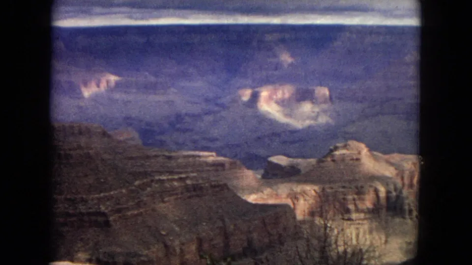 1967 8mm home-movie frame with foreground canyon cliffs, distant pale rock shelves, and blue-purple haze across the Grand Canyon.