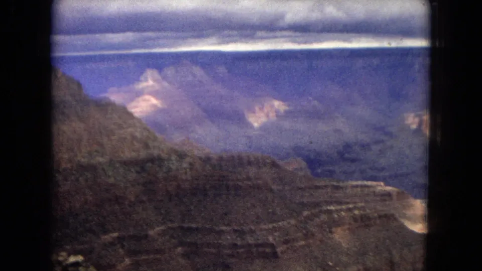 1967 8mm frame showing a Grand Canyon wall in the foreground with distant mesas and ravines receding into blue-purple atmospheric haze.