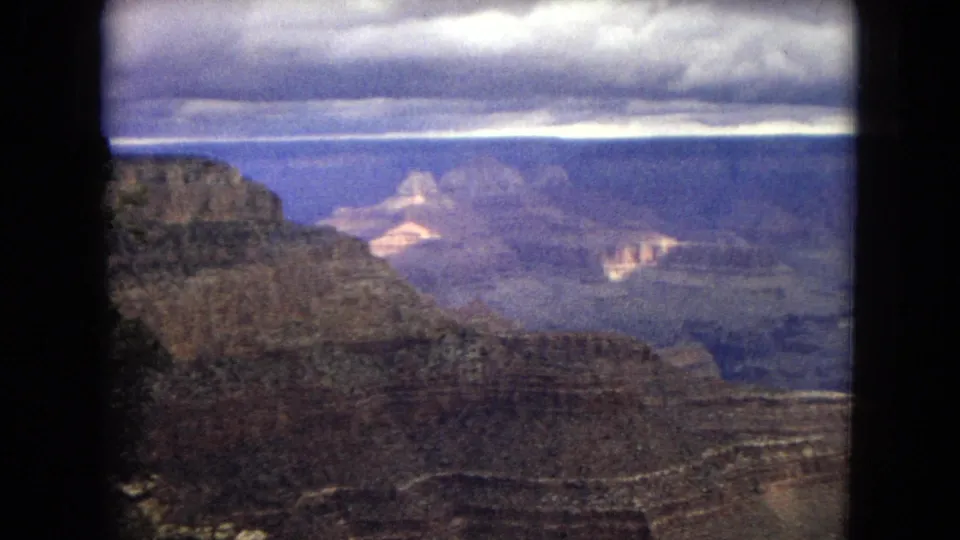 1967 8mm home-movie view of the Grand Canyon with layered rock walls, distant mesas, and a cloudy sky seen from a high overlook.