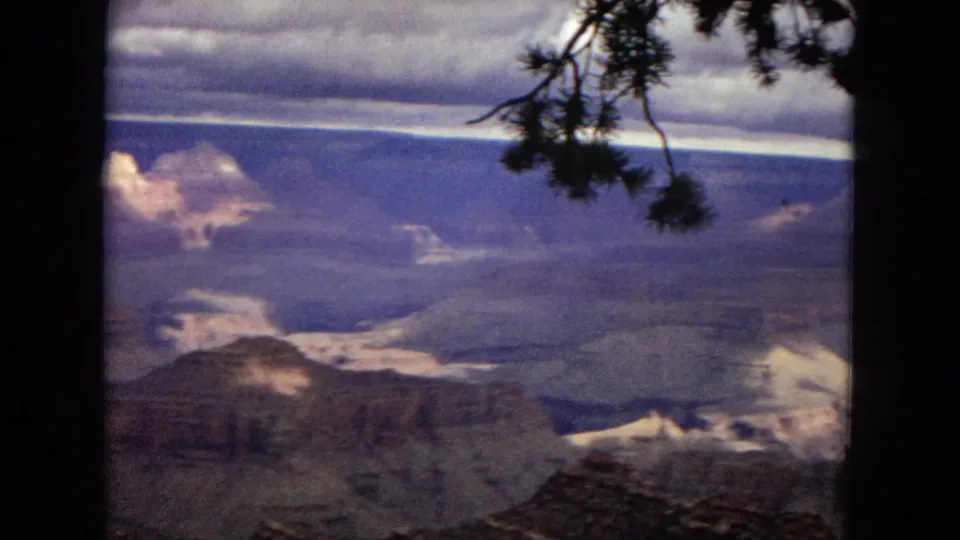 Grand Canyon home-movie frame from 1967 with dark pine branches in the foreground and distant layered canyon walls beneath a cloudy sky.