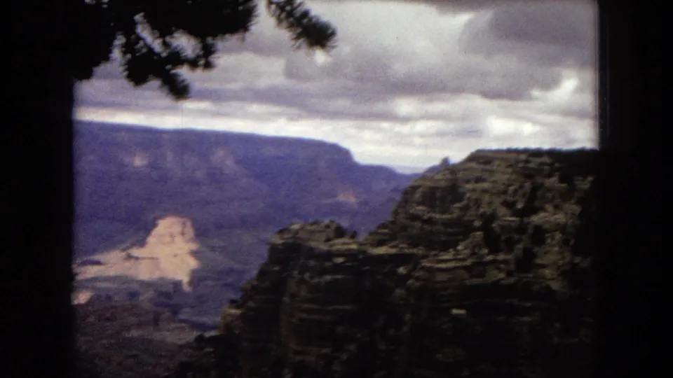 1967 8mm frame of the Grand Canyon from an overlook, showing a rocky foreground formation, distant canyon walls, and a heavy cloud layer above.