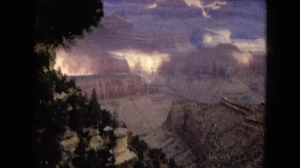 1967 home-movie frame looking across the Grand Canyon in Arizona, with dark tree silhouettes in the foreground and illuminated canyon formations under broken clouds.
