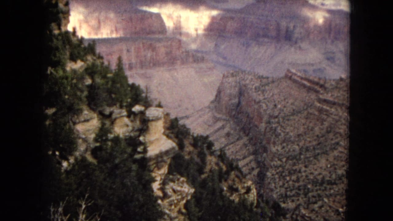 1967 Arizona 8mm frame looking from a pine-edged cliff across layered canyon walls and distant rock formations.
