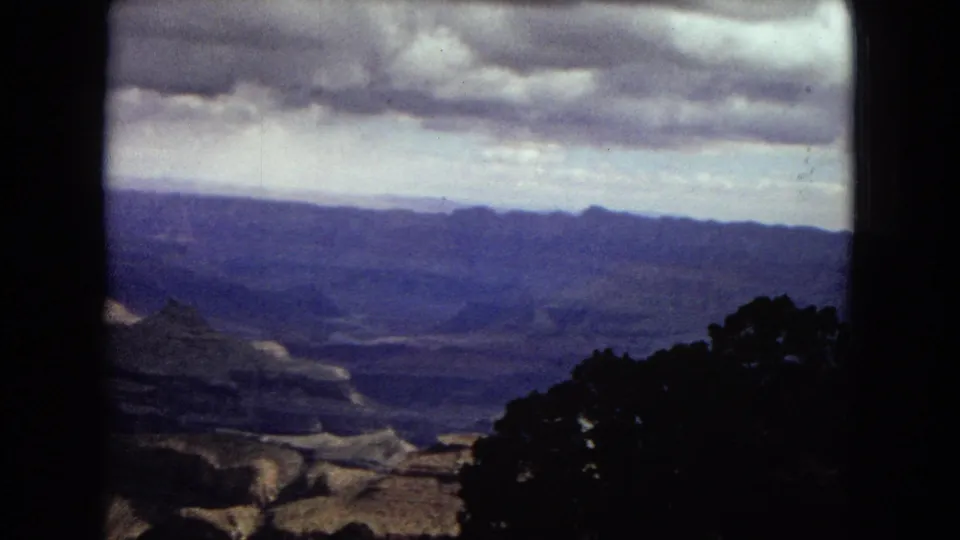 1967 Arizona 8mm panorama across a deep canyon landscape, with cloud cover above distant mesas and dark foreground trees.