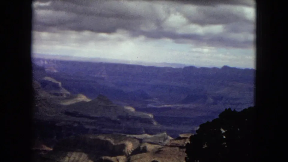 1967 Arizona home-movie frame showing an expansive canyon view beneath heavy clouds, with distant ridges and dark trees along the overlook.