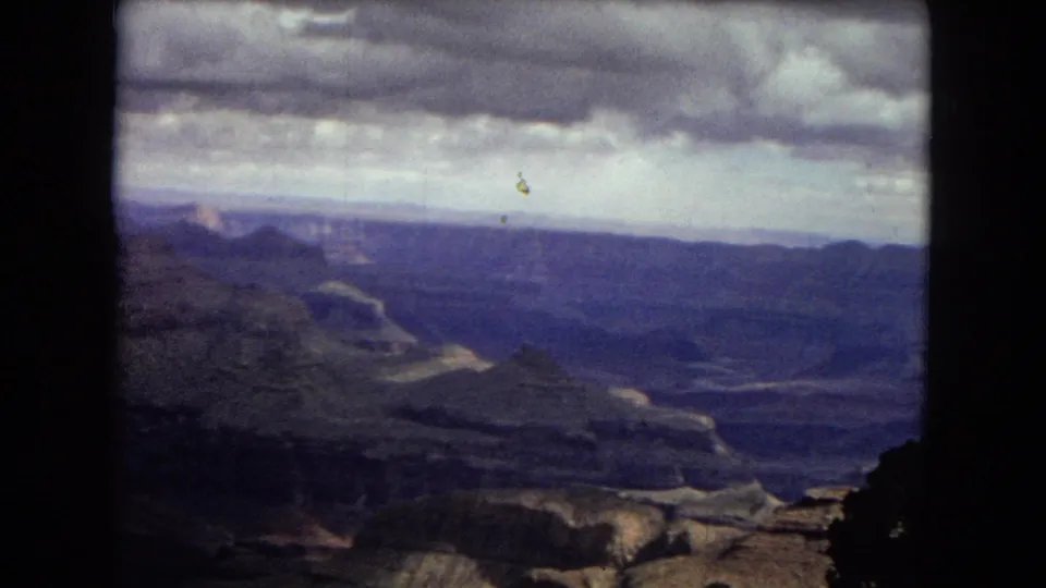 1967 8mm home-movie frame of the Grand Canyon in Arizona, showing layered cliffs, a broad valley, distant ridges, and a moody overcast sky.