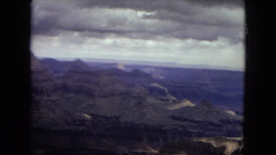 1967 8mm travel frame looking across the Grand Canyon, with distant mesas, deep blue-purple canyon layers, and a low ceiling of clouds.