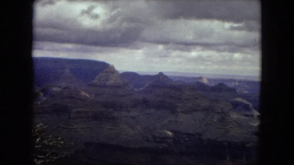 1967 8mm home-movie view of Grand Canyon ridges and valley layers, with soft focus, muted color, and a broad band of overcast sky.