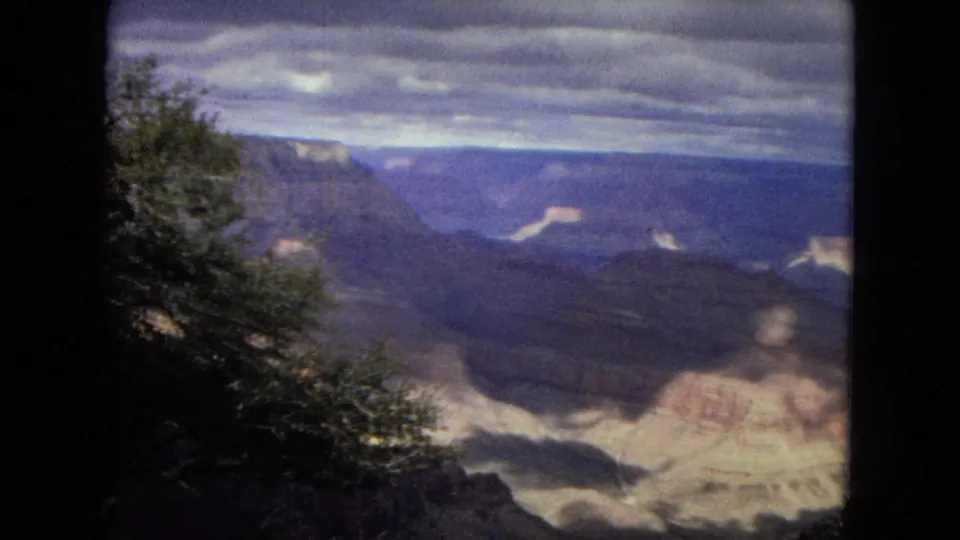 1967 8mm frame from an Arizona Grand Canyon overlook, showing foreground shrubs, steep canyon walls, pale rock layers, and dramatic cloud cover.
