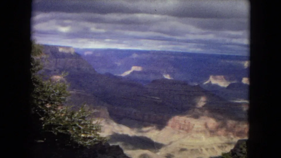 1967 8mm Arizona home-movie frame showing a Grand Canyon panorama beneath an overcast sky, with layered rock formations and foreground vegetation.