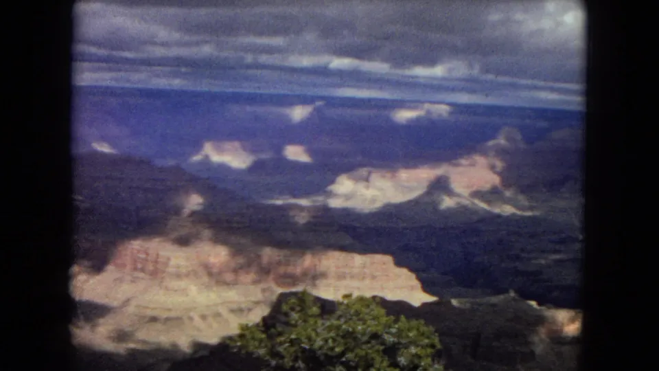 1967 8mm Arizona home-movie frame showing Grand Canyon ridges, sunlit pale rock shelves, and dense cloud cover across the horizon.