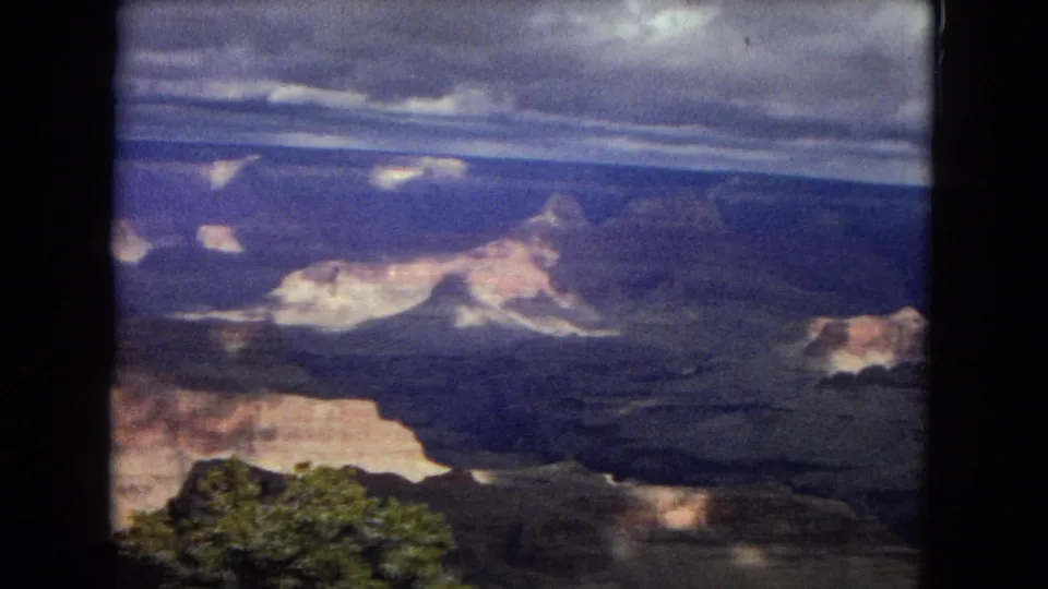 1967 8mm Arizona frame of a Grand Canyon overlook, showing dark canyon slopes, pale cliff faces, foreground brush, and a moody cloud layer.