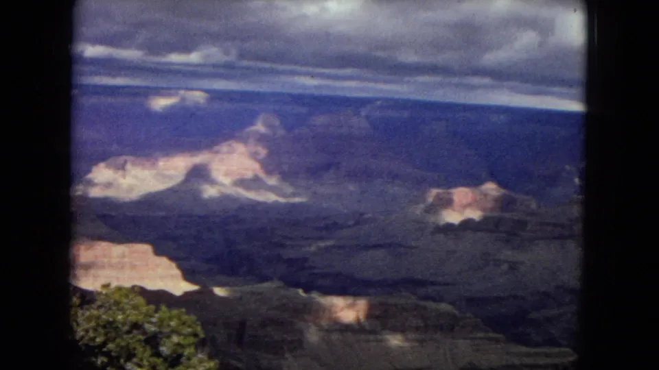 1967 8mm home-movie frame showing a broad Grand Canyon overlook with layered cliffs, blue shadows, clouds, and slight foreground vegetation.