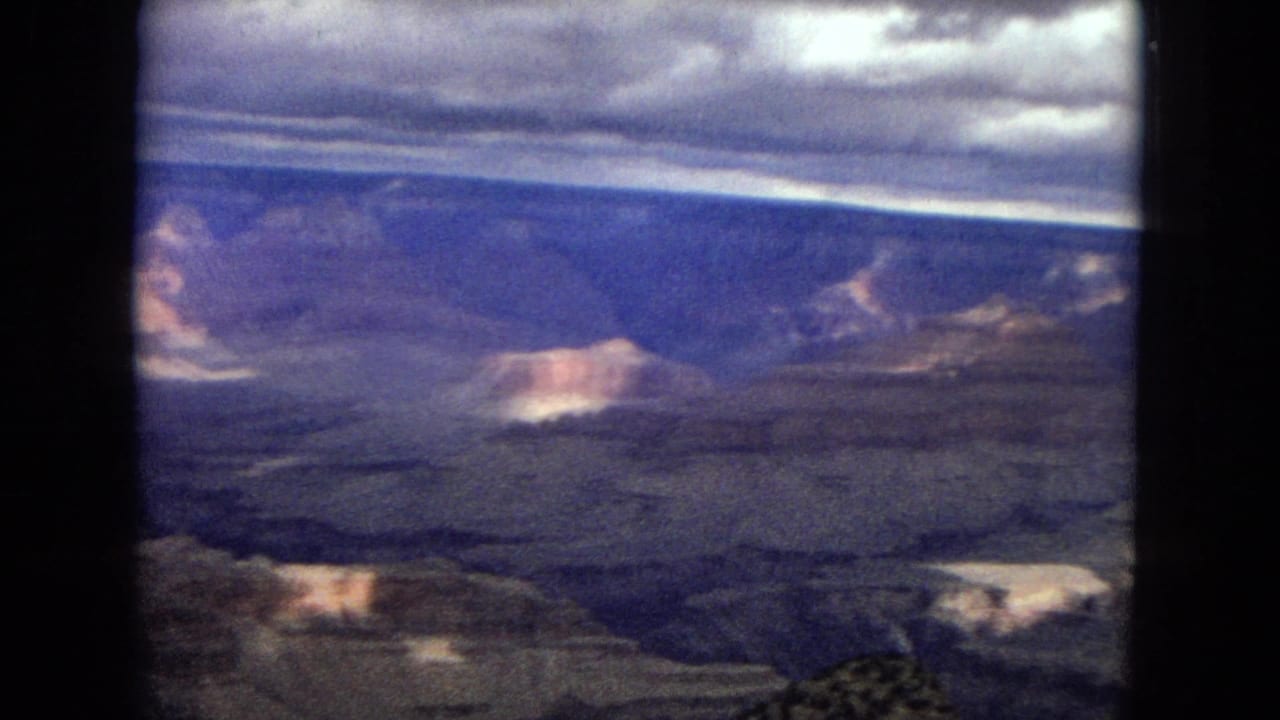 1967 Arizona home-movie frame with a distant Grand Canyon wall, heavy clouds, pale sunlit rock areas, and dark vertical film borders.
