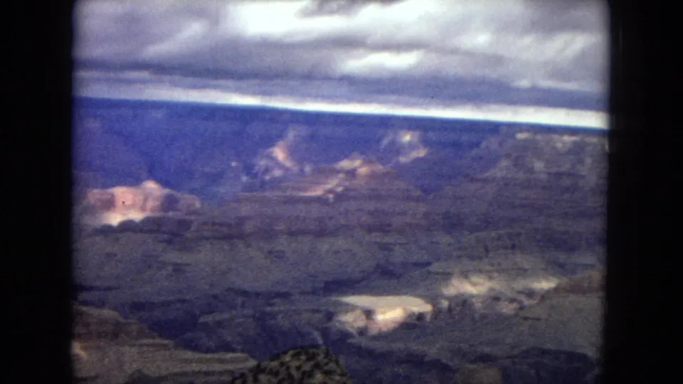 1967 8mm frame showing the Grand Canyon from an overlook, with layered rock, bright cloud breaks, dark ravines, and low vegetation at the bottom edge.
