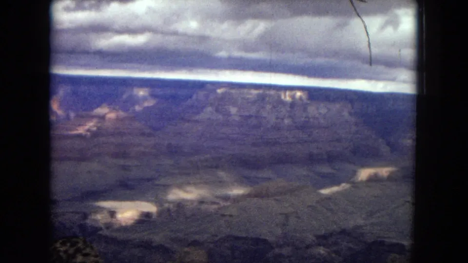 1967 Arizona home-movie frame of the Grand Canyon with broad rock layers, a bright cloud break near the horizon, dark film borders, and a thin scratch near the upper right.