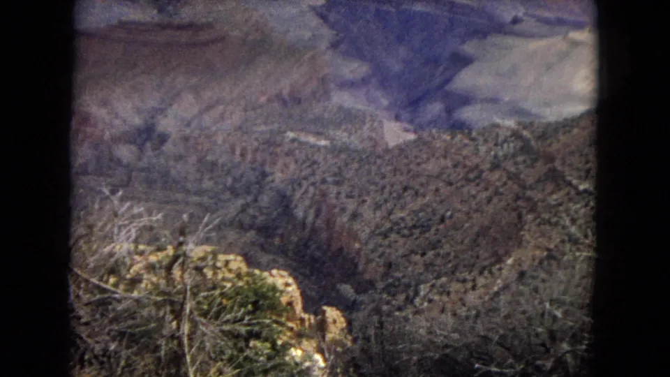 1967 8mm footage frame showing a Grand Canyon overlook view with scrubby foreground vegetation, rocky ledges, and layered canyon walls in the distance.