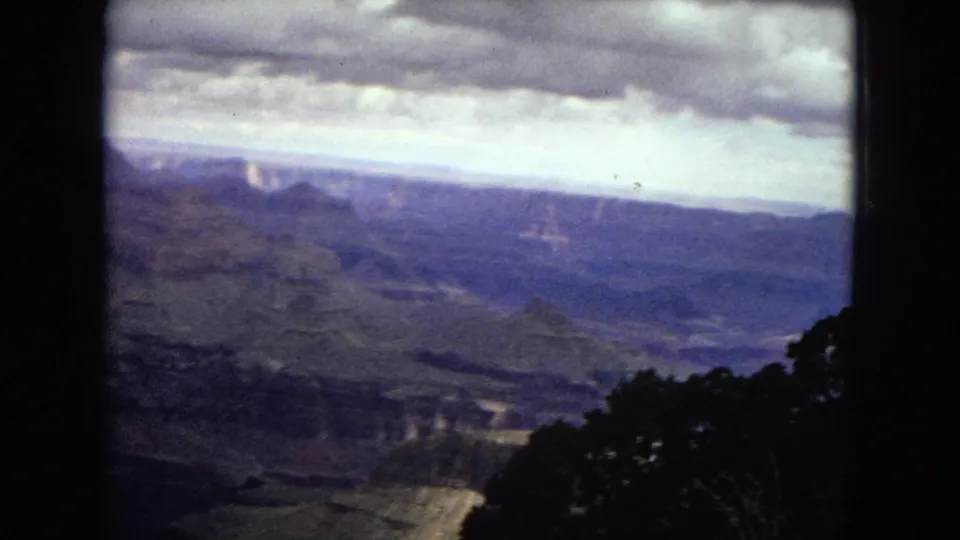 1967 8mm frame of the Grand Canyon from a high vantage point, with a clouded sky, distant rock formations, shadowed canyon floor, and dark foreground trees.