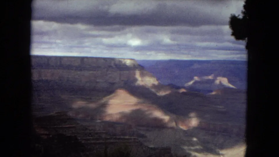 1967 8mm home-movie frame showing a broad Grand Canyon overlook, distant mesas, shadowed ravines, foreground brush, and a cloudy horizon.