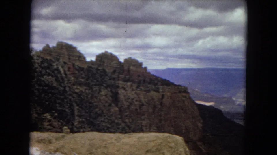1967 8mm view of a rugged Grand Canyon cliff face beside a stone overlook edge, with blue distance and heavy clouds across the horizon.