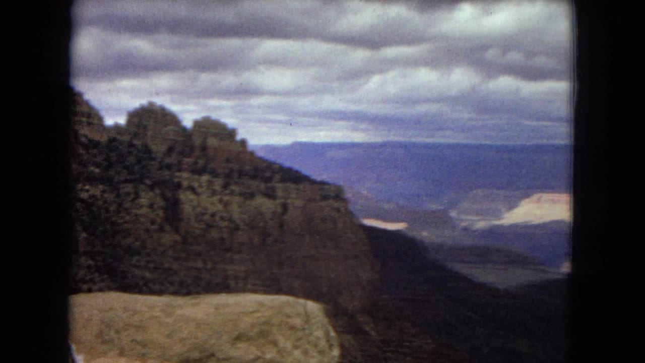 1967 8mm view across the Grand Canyon area from a high overlook, with shadowed canyon walls, distant blue mesas, heavy clouds, and a pale rock ledge in front.