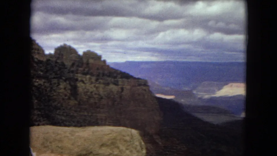 1967 8mm view across the Grand Canyon area from a high overlook, with shadowed canyon walls, distant blue mesas, heavy clouds, and a pale rock ledge in front.