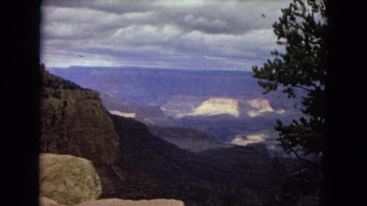 1967 8mm Grand Canyon vista with a cliff face on the left, pale sunlit formations in the distance, layered mesas, a cloudy sky, and a pine tree entering from the right edge.