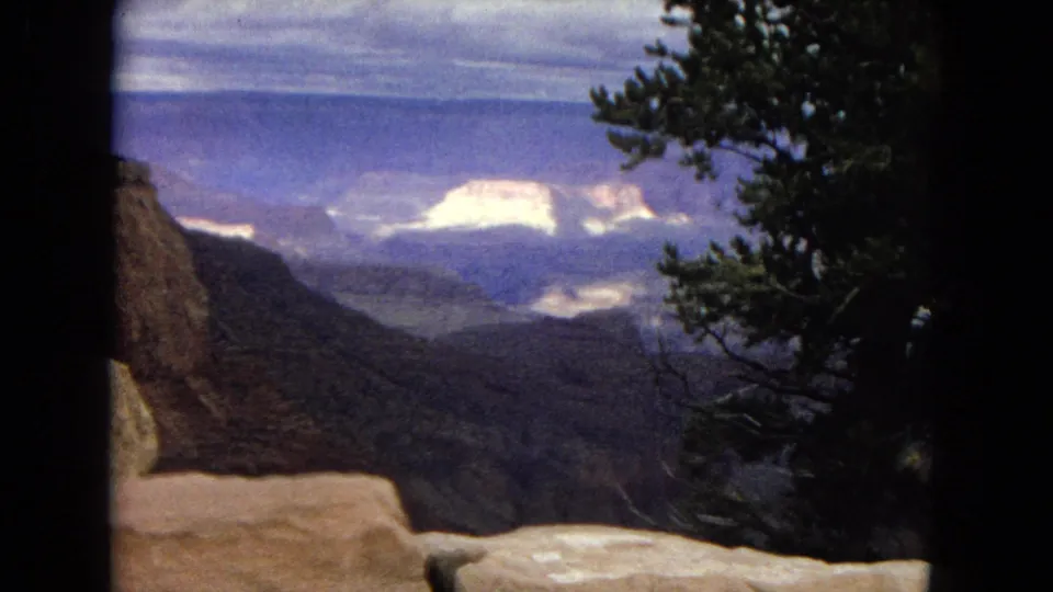 1967 8mm home-movie frame looking across the Grand Canyon landscape, with a foreground stone ledge, shadowed cliff wall, distant pale rock formations, and a dark pine tree on the right.