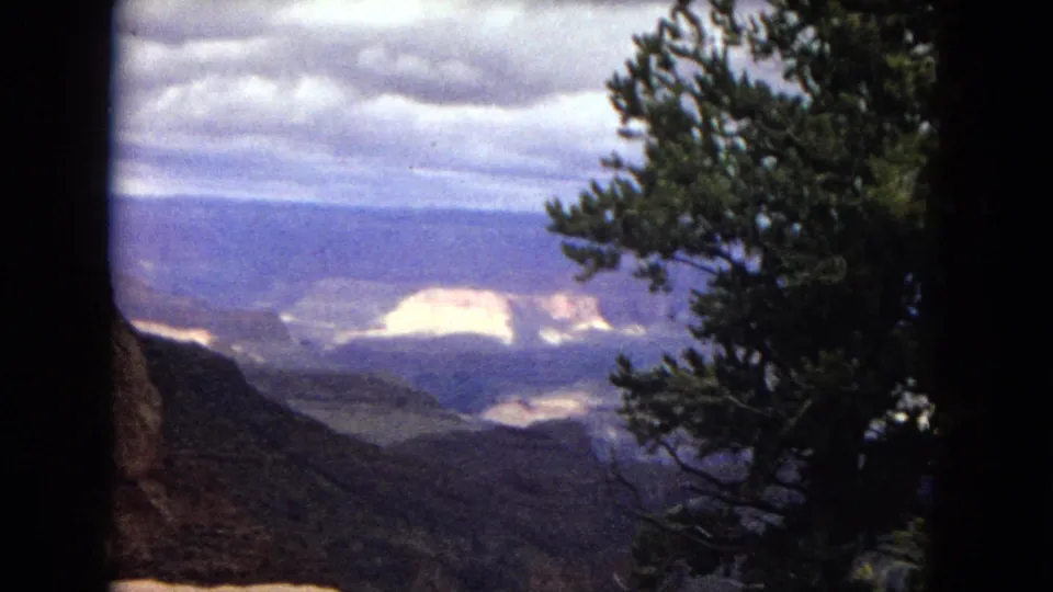 Final 1967 8mm frame of the Grand Canyon vista, with a pine tree framing the right side, dark canyon slopes below, pale distant formations, blue mesas, and a cloud-filled sky.