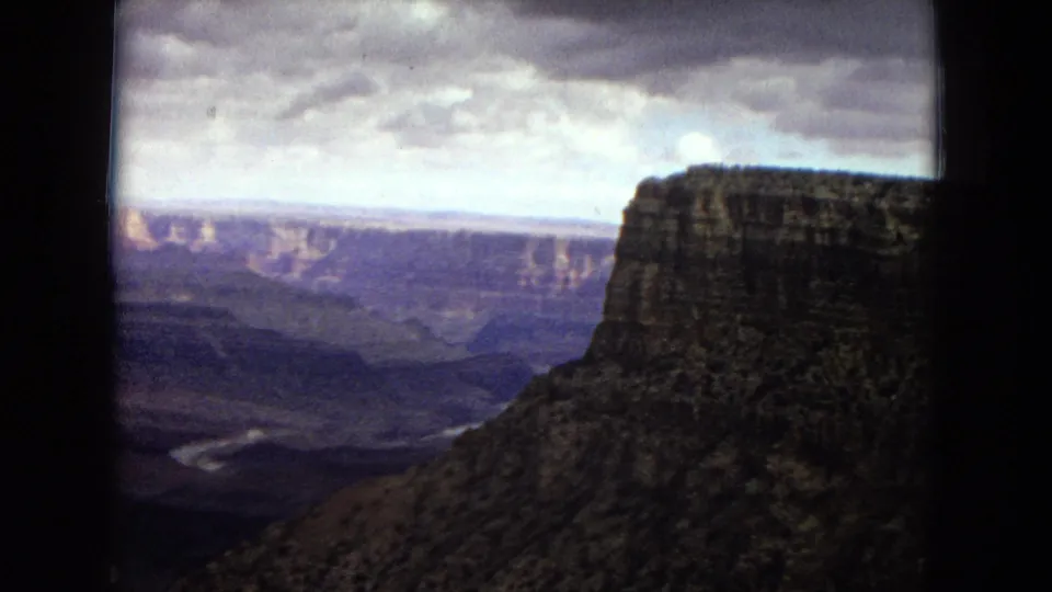 1967 8mm home-movie frame of the Grand Canyon in Arizona, showing a broad canyon view with layered rock formations, a distant river bend, cloudy sky, and dark rounded film borders.