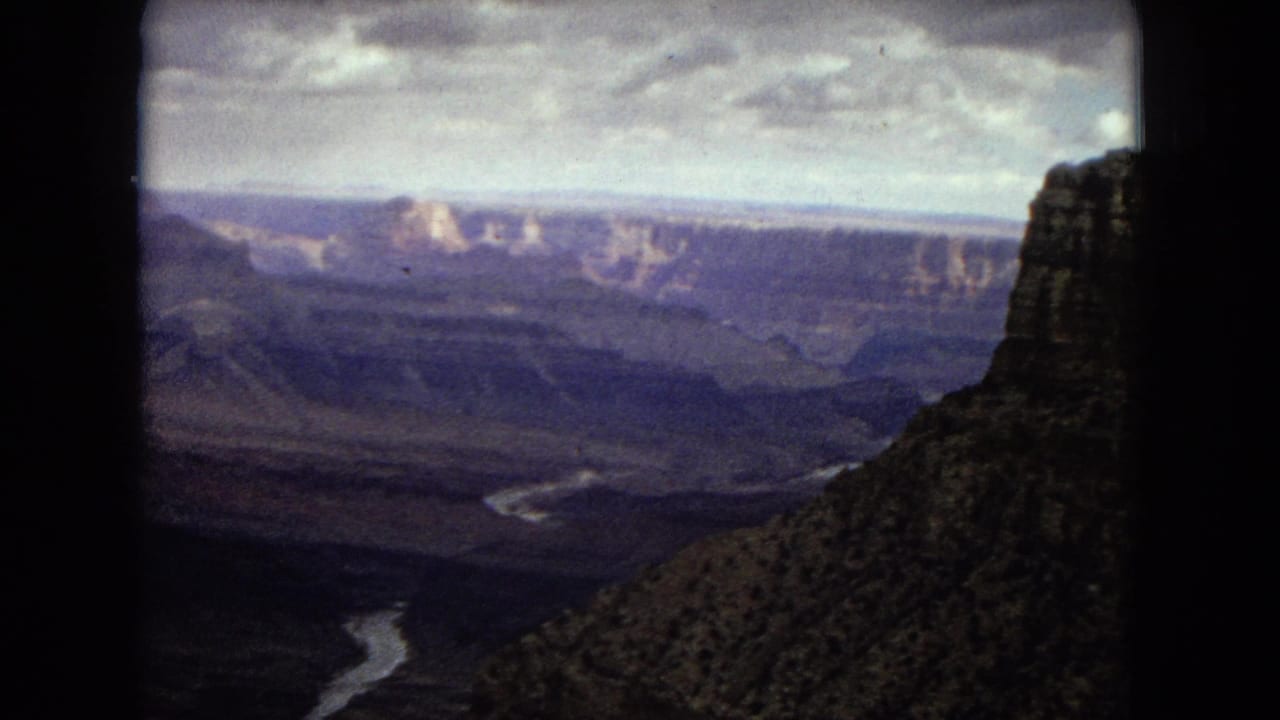 1967 8mm frame from an Arizona Grand Canyon panorama, with the Colorado River appearing as a pale winding line below distant layered cliffs and a dark film-frame edge at center.