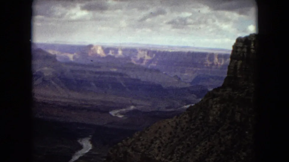 1967 8mm frame from an Arizona Grand Canyon panorama, with the Colorado River appearing as a pale winding line below distant layered cliffs and a dark film-frame edge at center.