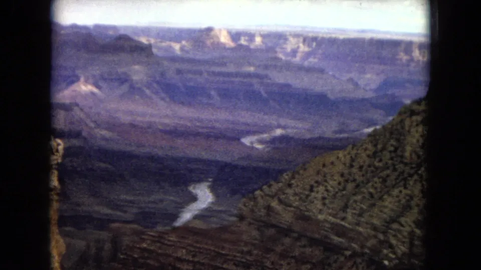 1967 8mm home-movie view of the Grand Canyon, looking across layered rock walls and a winding river far below with a foreground slope and soft, faded color.